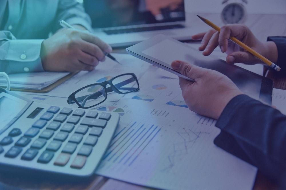 Business One Tax & Accounting professionals reviewing financial charts and reports at a desk, with a calculator, tablet, glasses, and paperwork — representing accounting, tax planning, and financial consulting services
